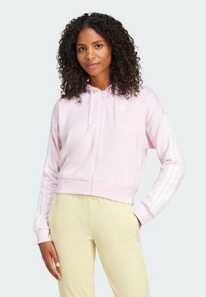 Rosa hettegenser med glidelås, hvite striper på ermene, og en liten logo. Båret med gule treningsbukser som viser en avslappet passform.