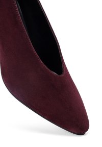 Chaussure à talon haut en daim bordeaux avec un bout pointu et un design découpé sur le côté, présentant une texture lisse et une semelle noire.