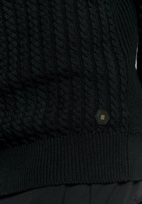 Zwarte gebreide trui met een kabelpatroon, een geribde zoom en een hexagonaal logo patch met gouden accenten aan de zijkant.