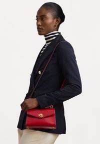 Κόκκινη δερμάτινη τσάντα ώμου με χρυσές λεπτομέρειες, σχεδίαση με καπάκι και διακριτό λογότυπο. Συνδυάζεται με ναυτικό σακάκι και ριγέ ψηλό λαιμό.