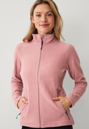 Młoda kobieta w różowej bluzie polarowej na zamek z rękami w kieszeniach, stojąca na tle jednolitego jasnoszarego tła.