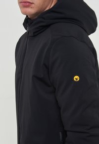 Giacca nera con cappuccio, tessuto texturizzato, chiusura con zip e un logo giallo sulla manica. Presenta una vestibilità slim e tasche laterali.