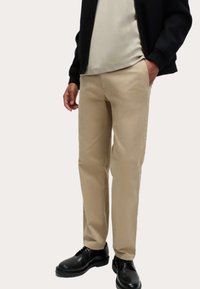 Beige broeken met een rechte pasvorm, gemaakt van een soepele katoenstof en met zijzakken, gecombineerd met zwarte schoenen en een donker vest met ritssluiting.