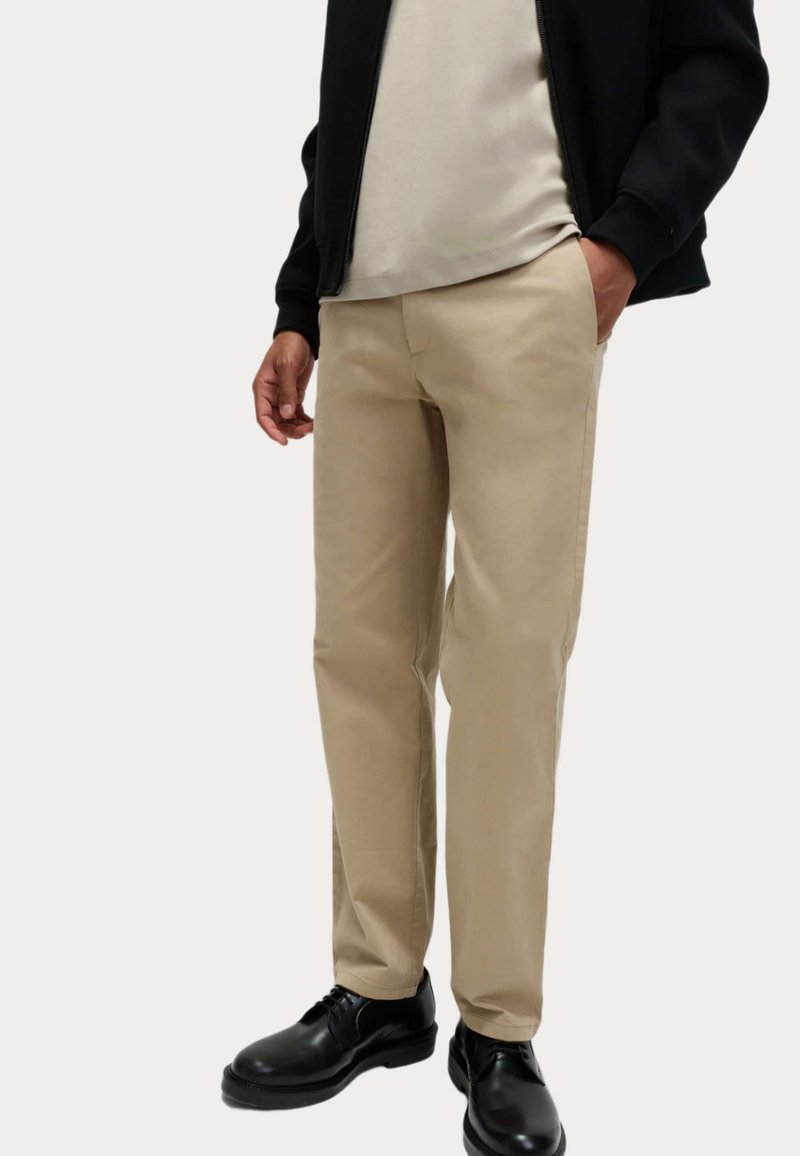 Beige broeken met een rechte pasvorm, gemaakt van een soepele katoenstof en met zijzakken, gecombineerd met zwarte schoenen en een donker vest met ritssluiting.