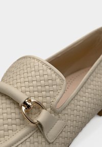 Μπεζ υφασμένα δερμάτινα μοκασίνια με μικρό διακοσμητικό φιόγκο και χρυσούς μεταλλικούς λεπτομέρειες. Η εσωτερική πλευρά είναι λεία με διακριτικές τρύπες.