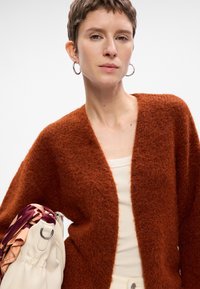 Rustfarvet, fluffy cardigan med åben front, der har en afslappet pasform. Sammen med en cremefarvet håndtaske og et mønstret tørklæde.