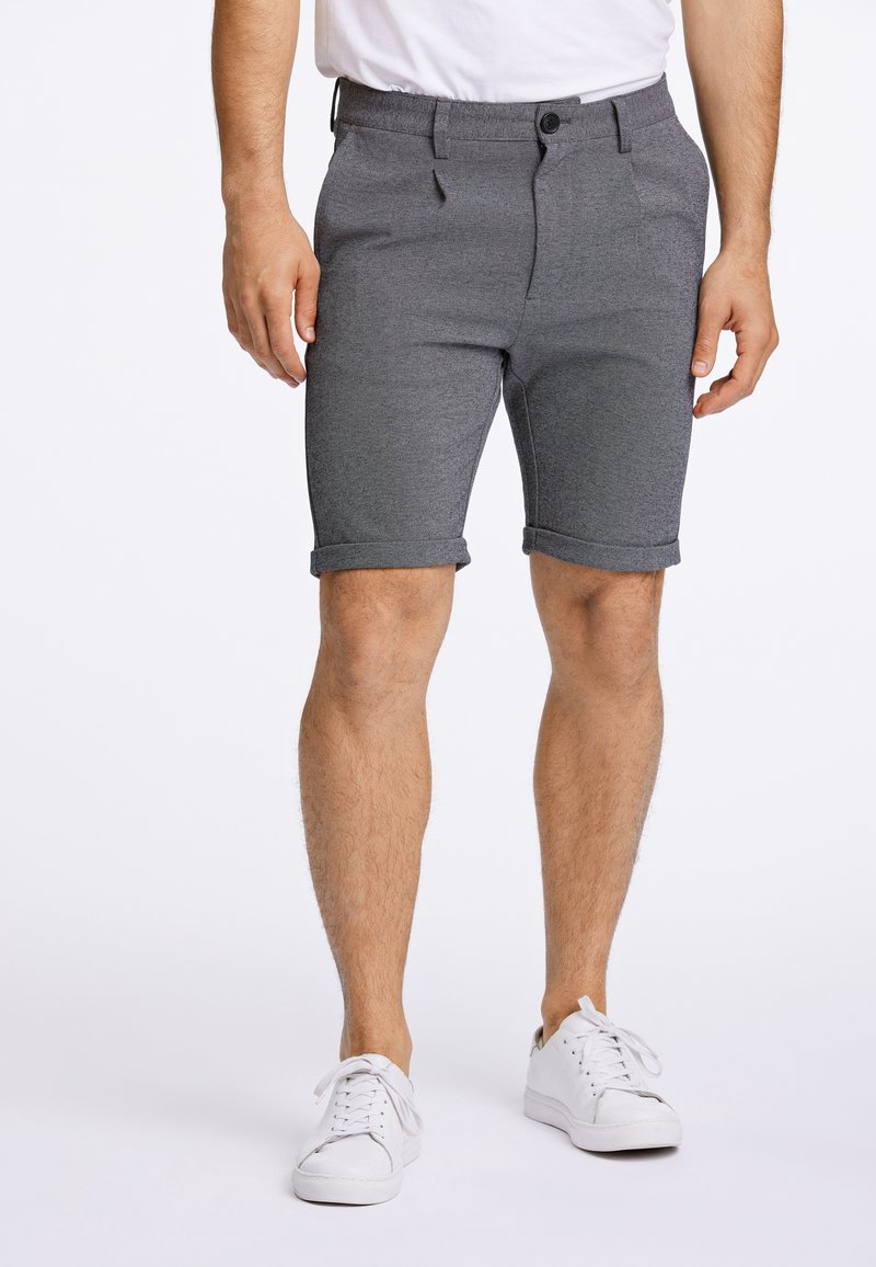 Homme portant un short gris sur mesure avec ourlets retroussés, un t-shirt blanc et des baskets blanches, debout sur un fond blanc.