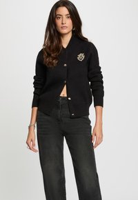Cárdigan negro hecho de material suave, con un cierre frontal con botones dorados y un emblema decorativo en el pecho. Puños acanalados.