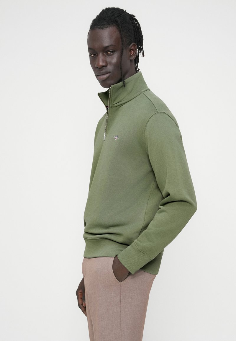 Sudadera verde con cremallera en un tejido texturizado, que presenta un cuello alto y un pequeño logo. Combinada con pantalones marrón claro de corte ajustado.