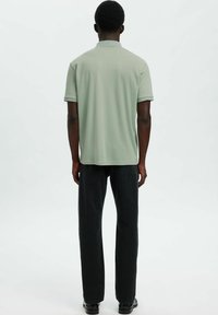 Svetlo zelena polo majica z ovratnikom in kratkimi rokavi, kombinirana s črnimi hlačami z ravnimi hlačnicami in črnimi čevlji, viden od zadaj.