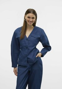 Giacca di jeans con maniche lunghe, scollo a V e due tasche sul petto, con grandi bottoni e una vestibilità comoda, in tessuto blu scuro.