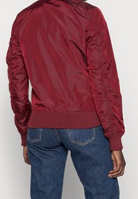 Burgundy bomberjacka i glänsande nylonmaterial, med ribbade muddar och nederkant samt samlade ärmdetaljer. Matchad med blå denimjeans.