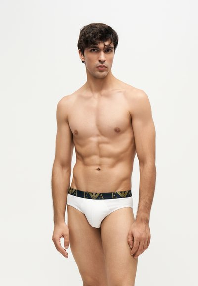 Jeune homme tonique aux cheveux foncés portant un slip blanc avec une ceinture noire et dorée brandée, debout sur un fond clair uni.