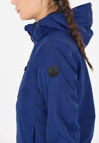 Marineblauer Kapuzenjacke mit matter Oberfläche, ausgestattet mit einem schwarzen, runden Logo am Ärmel und strukturierten Nähten. Verstellbare Kapuze mit Druckknopfverschlüssen.