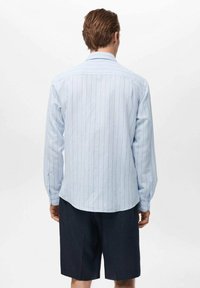 Camisa de manga larga azul clara con rayas verticales, puños abotonados y dobladillo curvado, combinada con pantalones cortos oscuros. Fondo liso.