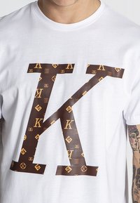 Bílý bavlněný tričko s velkým hnědým logem ve tvaru písmene "K" na přední straně. Hladký materiál, krátké rukávy.
