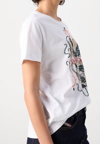 Bílý bavlněný tričko s krátkým rukávem, s barevným vyšívaným designem na přední straně. Nosené s tmavými džíny.
