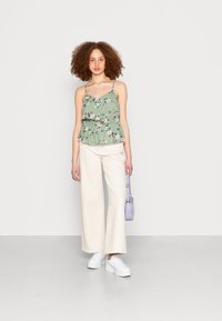 Top peplum floral verde claro com alças finas, combinado com calças largas brancas e ténis brancos de plataforma. Segura uma pequena bolsa roxa.