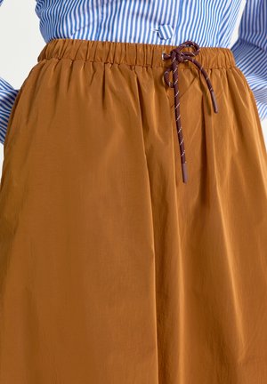 Fustă maro cu talie elastică și șnur, purtată cu o cămășă în dungi albastre și albe băgată în fustă.