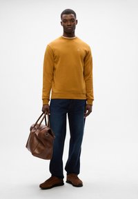 Felpa gialla senape con polsini a coste, jeans in denim blu, borsa in pelle marrone e scarpe in suede marroni; design pulito e minimal.