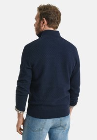 Navy-musterter Sweatshirt mit hohem Kragen, gerippten Bündchen und Saum, kombiniert mit hellblauen Jeans. Die Oberfläche weist ein waffleartiges Design auf.