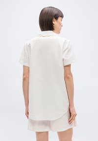 Femme aux cheveux raides et foncés portant une chemise crème ample à manches courtes et un short assorti, debout devant un fond clair uni.