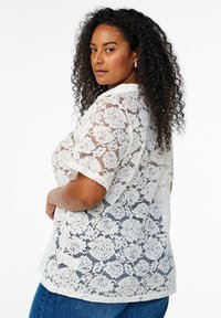 Chemise en dentelle blanche avec des motifs floraux et des manches courtes, superposée sur une base unie, au fit décontracté et avec des détails de col.
