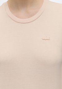 T-shirt beige à col rond en gros plan avec un petit logo crocodile ton sur ton brodé sur la partie gauche de la poitrine.