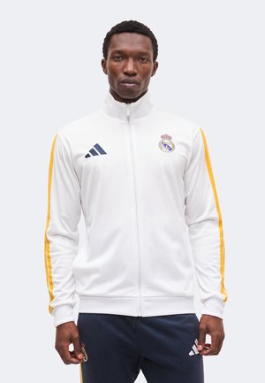Bărbat purtând o geacă albă cu fermoar și pantaloni bleumarin cu dungi galbene și siglele Real Madrid și Adidas, stând în fața unui fundal simplu.