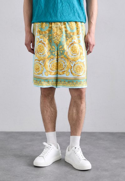 Turquoise shorts met ingewikkelde gouden bloemmotieven, relaxte pasvorm, boven de knie lengte, gecombineerd met witte sneakers en sokken.