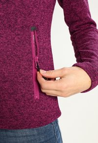 Fleece-paita syvää pinkkiä, teksturoitua kangasta, jossa on vetoketjullinen tasku, erottuvalla mustalla vetoketjun vetimellä ja istuvalla mitoituksella.