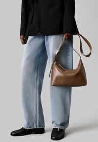 Brun læderhandbag med en buet form og lynlås, holdt af en person iført en sort blazer og lyseblå løse jeans.