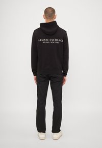 Sweat-shirt noir à capuche avec le logo "ARMANI EXCHANGE MILANO / NEW YORK" en blanc dans le dos, porté avec un pantalon noir et des baskets blanches.