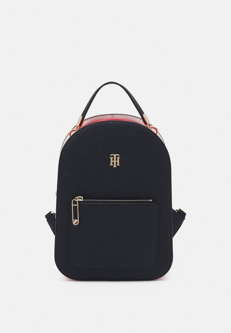 Petit sac à dos en cuir noir avec poignée supérieure, poche avant zippée avec fermeture éclair dorée et initiales du logo dorées sur une surface lisse texturée.