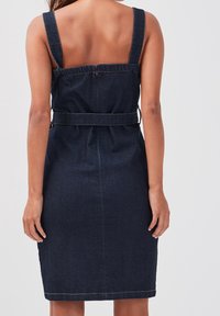 Tmavě modré džínové šaty se širokými ramínky, zúženým pasem a přiléhavým střihem. Mají hladkou strukturu a minimální šití. Pohled zezadu.
