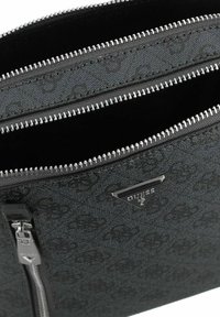 Bolso de piel sintética negra con compartimentos con cremallera, que presenta un sutil patrón de logo en relieve y detalles en hardware plateado. Etiqueta de logo en forma de triángulo exhibida.