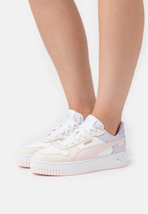 Pieds portant des baskets Puma blanches et rose pâle avec des semelles épaisses, un logo latéral et des lacets blancs sur un fond blanc uni.