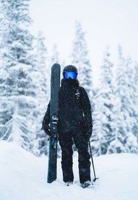Sprzęt narciarski przedstawiony z czarnym, ocieplanym kombinezonem i kaskiem. W tle zaśnieżony krajobraz z drzewami i padającym śniegiem.