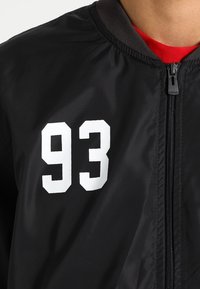 Schwarze Bomberjacke mit glatter Textur, ausgestattet mit der weißen Ziffer "93" auf der Brust und einem Reißverschluss vorne. Grauer gerippter Kragen.