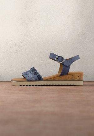 Blå kile-sandal med blomstret rem, ankelspænde, korkhæl og rillede beige sål på træflade mod hvid tekstureret væg.