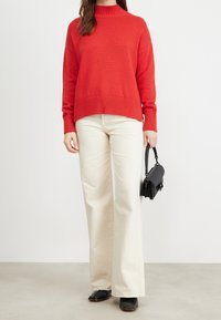 Rode gebreide trui met een hoge halslijn, geribbelde mouwen en zoom, gecombineerd met beige broek met wijde pijpen en een kleine zwarte handtas.