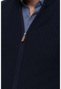 Marinblå zip-jacka med en texturerad våffelstickad mönster, avslutad med en silverfärgad dragkedja och en läderdragflik. Bärs över en ljusblå skjorta.