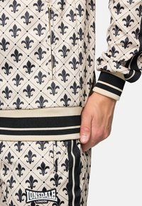 Beige tracksuit with a black fleur-de-lis pattern. Features black stripes on cuffs and waistband, with a zip pocket detail. Lonsdale logo visible.