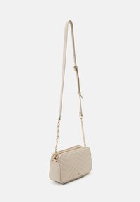 Quiltet beige crossover-veske med gullkjede-rem, med glidelås på toppen og chevronmønster, laget av glatt teksturert materiale.