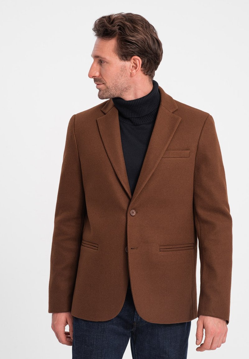 Brun ullblazer med klippta slag, enkel knappstängning och två framfickor, lagd över en svart polotröja. Robust textur, skräddarsydd passform.