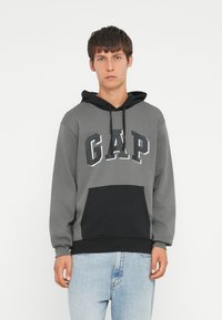 Hanorac gri cu accente negre, având logo-ul "GAP" în alb. Include o glugă și un buzunar frontal tip kangaroo, fabricat dintr-un material moale.