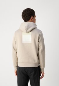 Bež dukserica s kapuljačom, meke teksture, s pravokutnom oznakom na leđima koja prikazuje naziv branda "Calvin Klein."