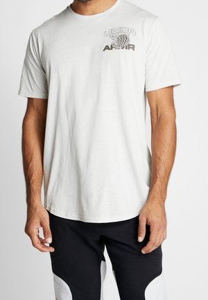 Mann trägt weißes Kurzarm-T-Shirt mit schwarzem "UNDR ARMOR" Schriftzug und Basketballkorb-Grafik auf der Brust, kombiniert mit schwarzen Hosen.