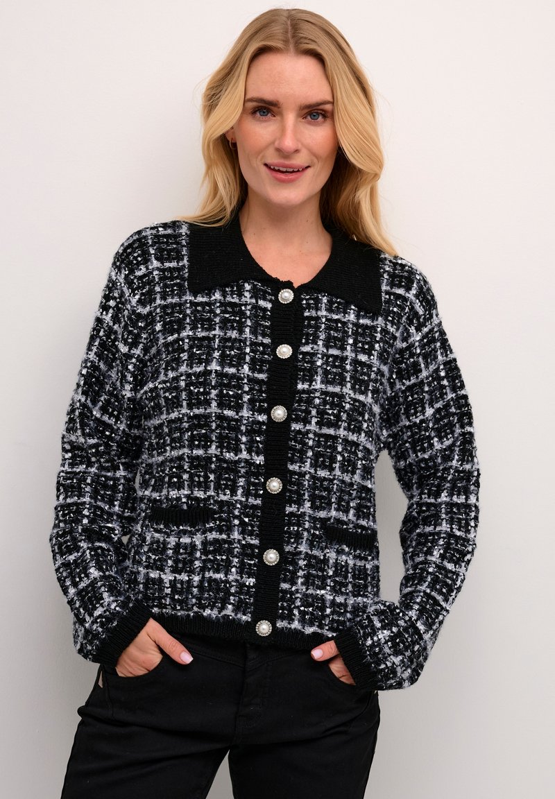Melnā un baltā šaha raksta kardigāns ar apkakli, lieli pērļu pogas, teksturēts audums un melni apdari pie aprocēm un apakšmalas.