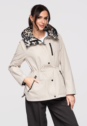 Donna che indossa una giacca beige con interno cappuccio in stampa leopardata, tasche con cerniera nere e pantaloni neri, posando con una mano in tasca.